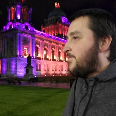 Jordan Upton outdoors at night, casual. Belfast City Hall lit purple behind.